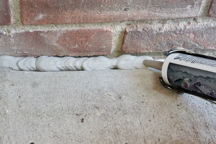 Applying sealant between a brick wall and concrete surface using a caulking gun.