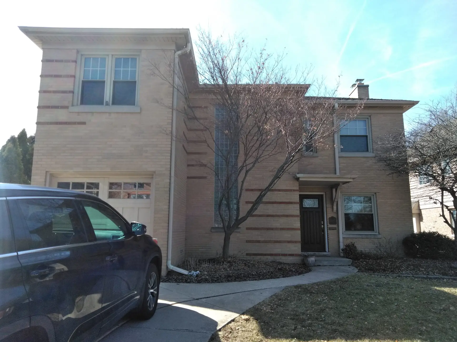 A two-story beige brick house with decorative brick patterns, a garage, a front yard with trees, and a parked dark-blue SUV in the driveway.