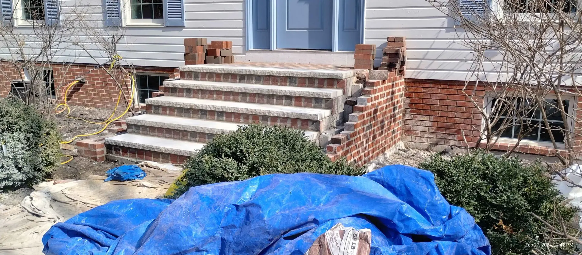 A front porch with brick steps under renovation, partially dismantled brickwork, construction tools, and blue tarps covering materials.