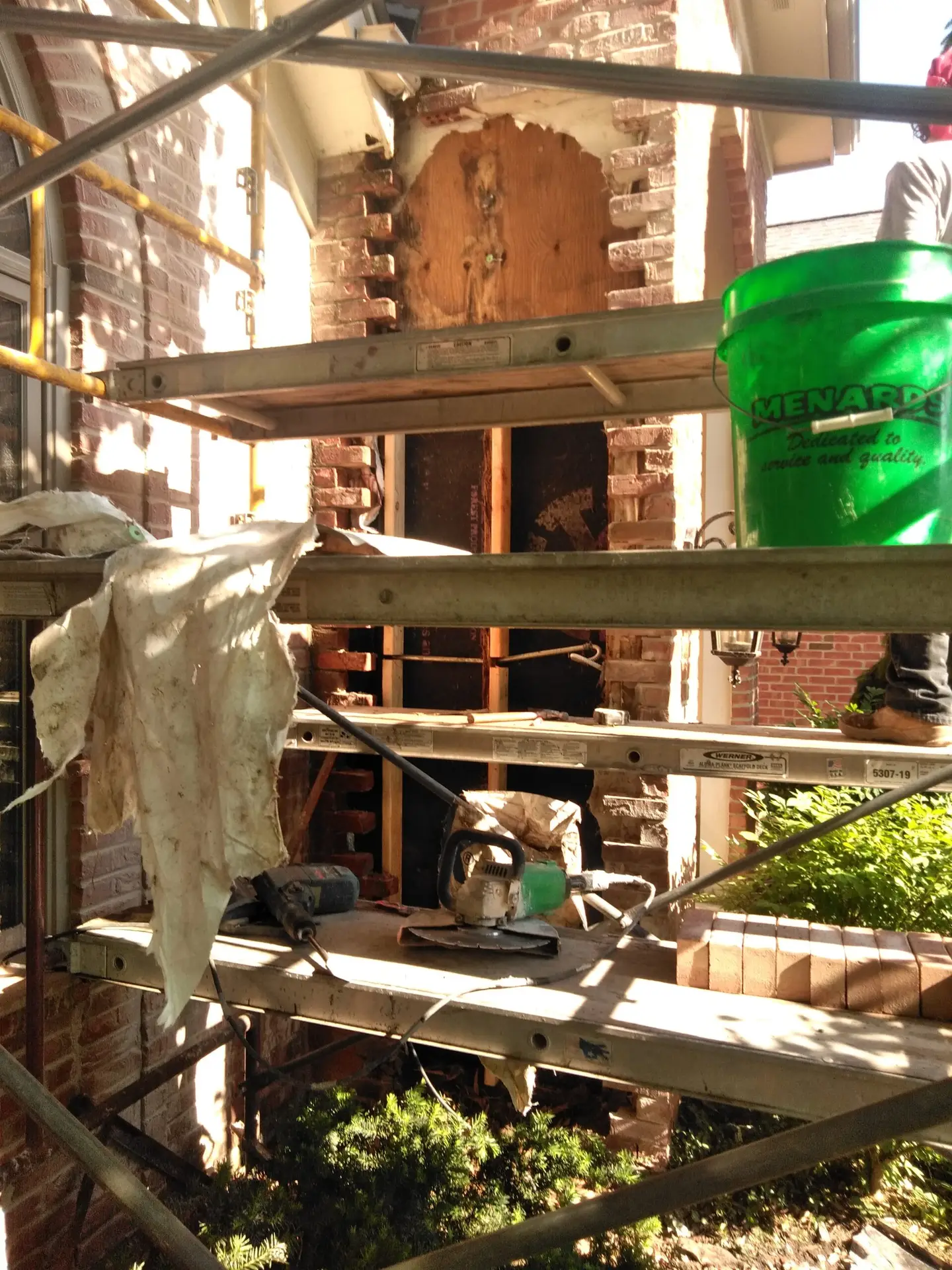 Construction scaffolding set up around a partially deconstructed brick wall, with tools, a green bucket, and a wooden panel covering the wall opening.